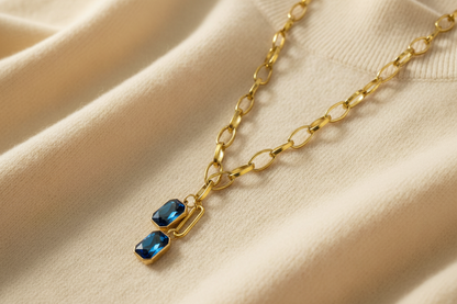 Gold chain necklace with sapphire blue double drop crystal pendant against and ivory sweater material background