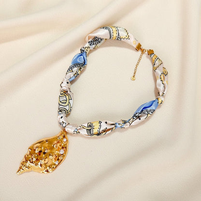 a scarf necklace with gold conch shell pendant on an ivory fabric background