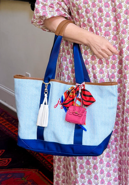 Person holding a blue and white tote bag with scarf twillie, leather tassel, and mini handbag charm in an indoor setting.