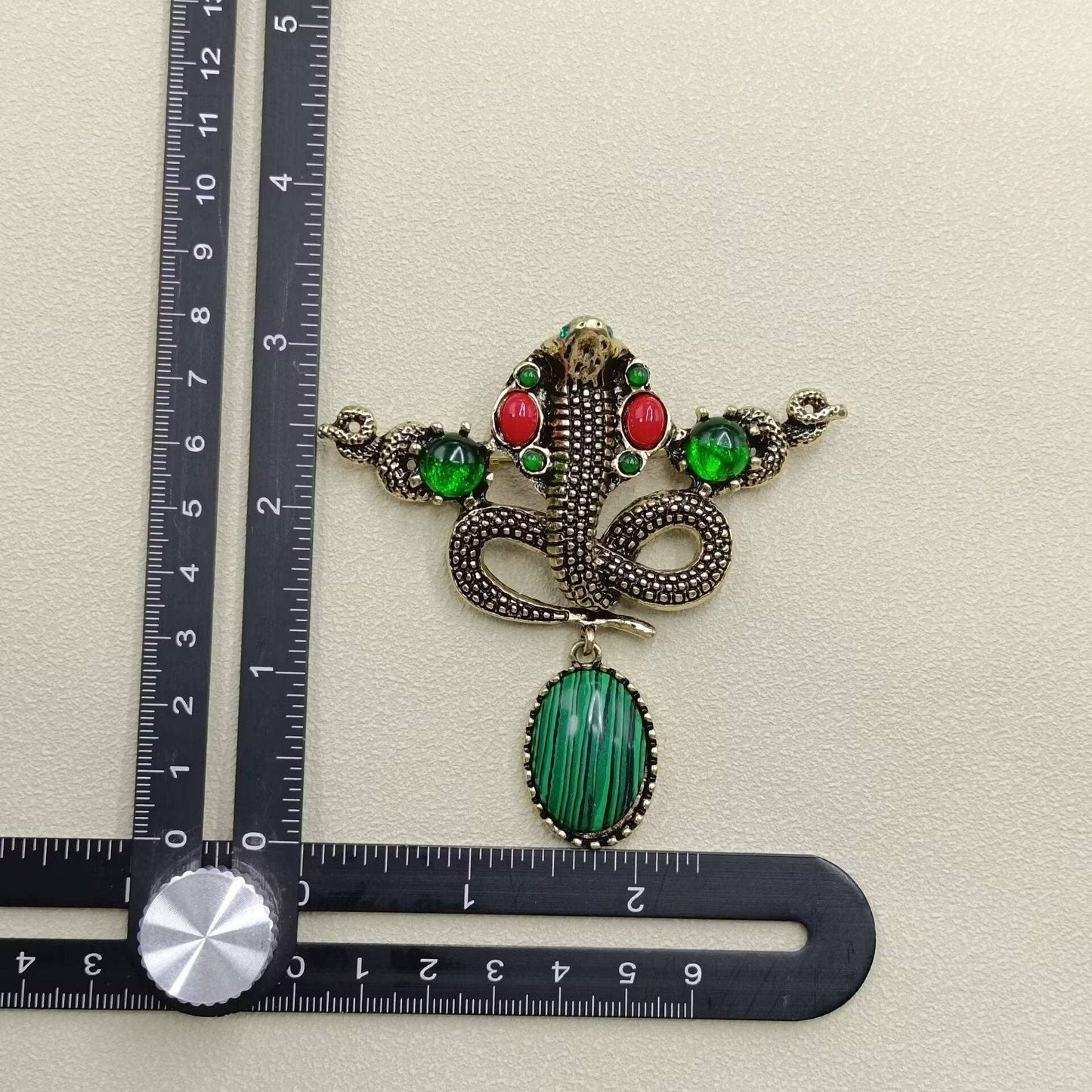 Decorative pendant with green stone and red accents next to a ruler on a beige background