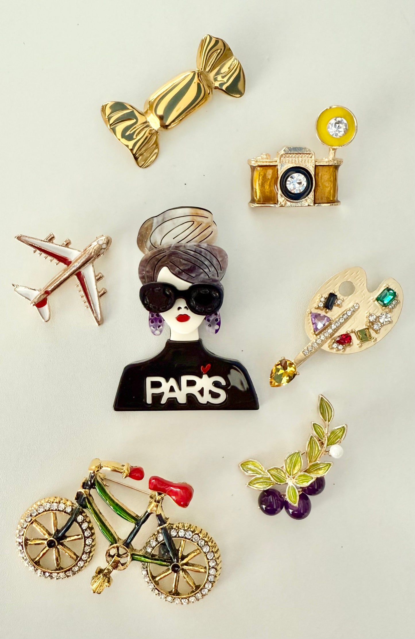 Parisian brooch collection showing fashionista, camera, palette, bicycle, airplane, candy, and grapes on a light background