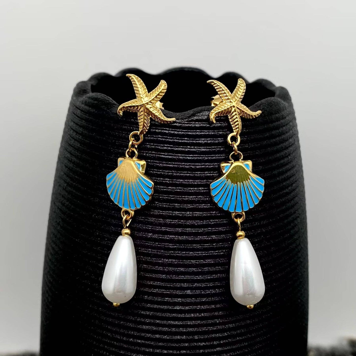 a pair of coastal inspired earrings with gold starfish and enamel clam with pearl drop against a black vase