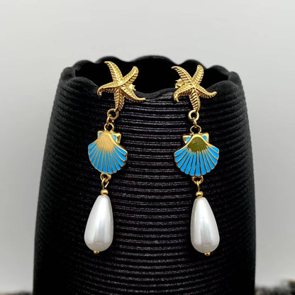 a pair of coastal inspired earrings with gold starfish and enamel clam with pearl drop against a black vase