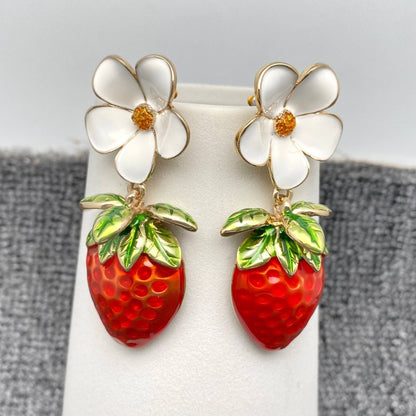 a pair of strawberry and daisy themed enamel earrings hanging from a white vase