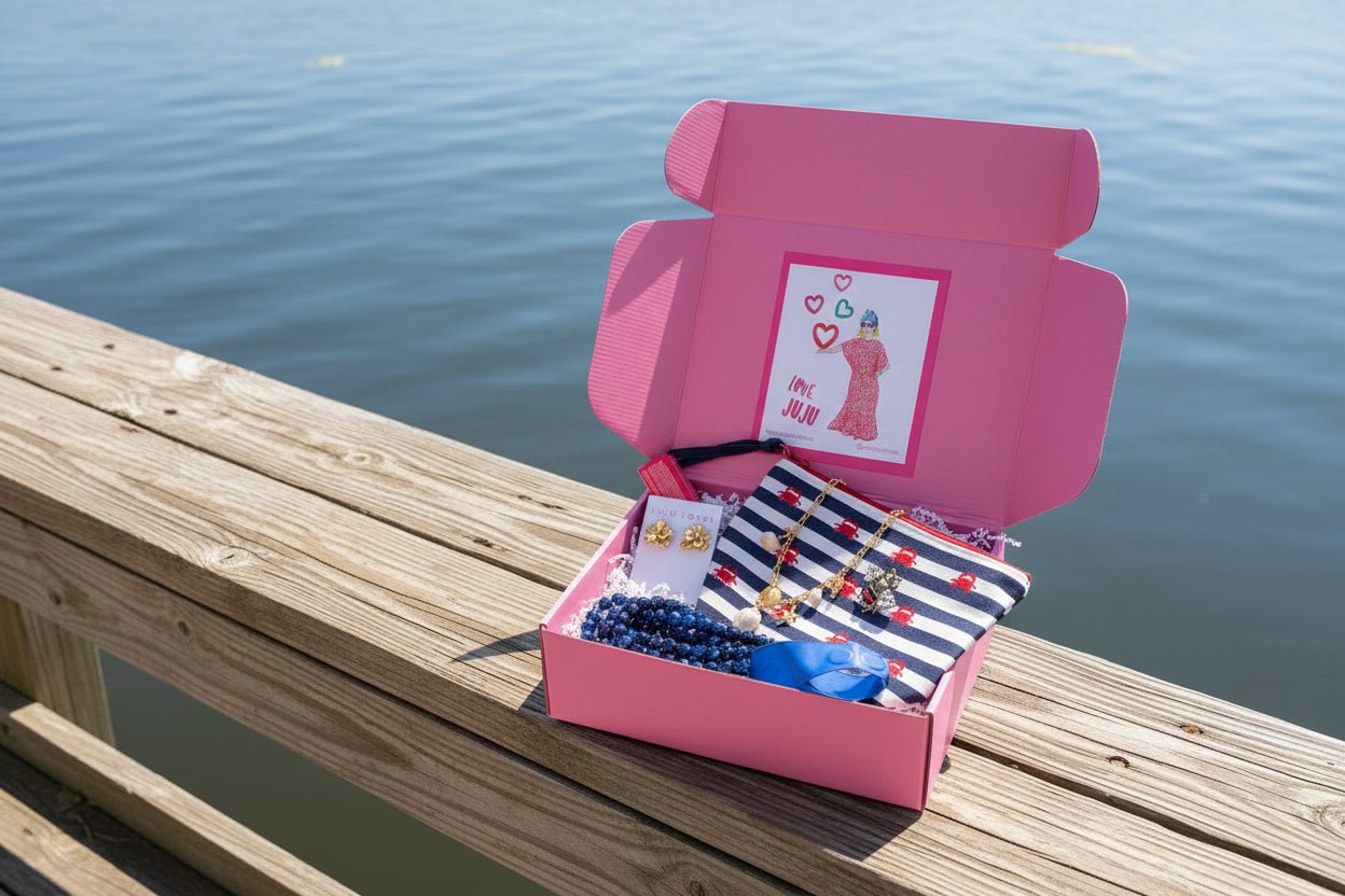 Pink Surprise Me JuJu Gift Box with necklaces, earrings, a pouch, and a brooch sitting on a dock with the water in the background.