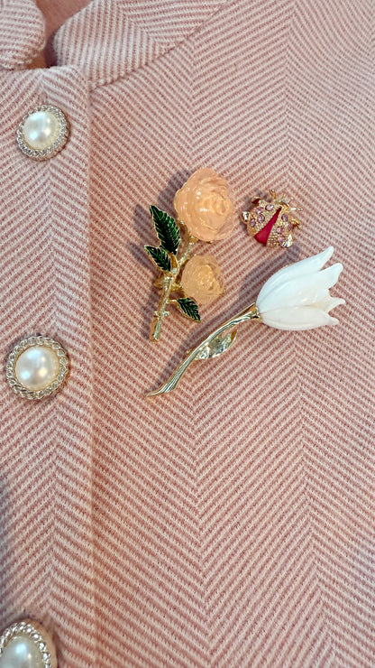 Close-up of 3 decorative brooches on a pink textured fabric background including a rose, tulip, and bee