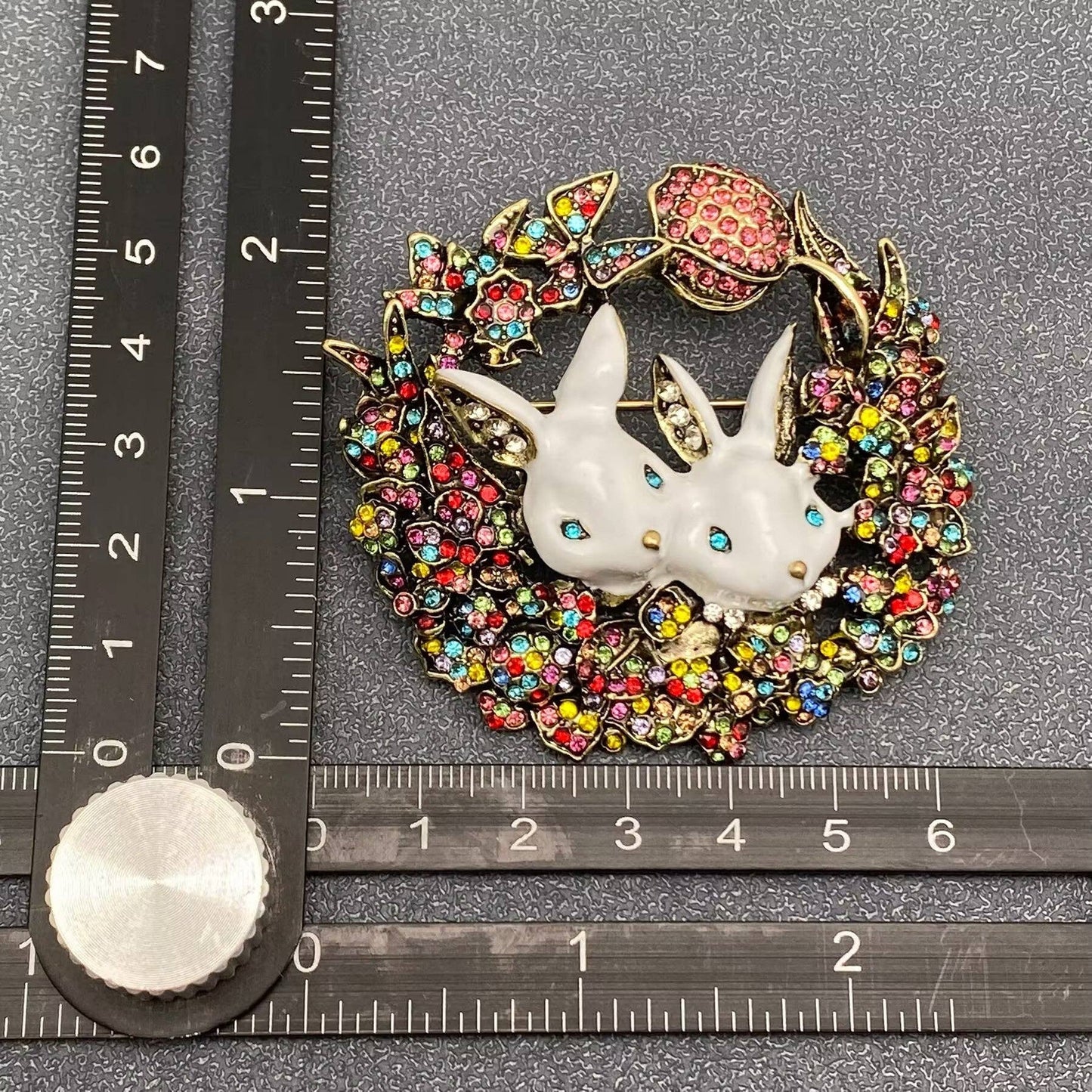 Decorative brooch with bunny design and a colorful gemstones wreath and a ruler for scale on a gray background