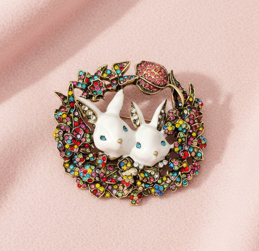 Brooch with two white rabbits in a colorful wreath on a pale pink fabric background