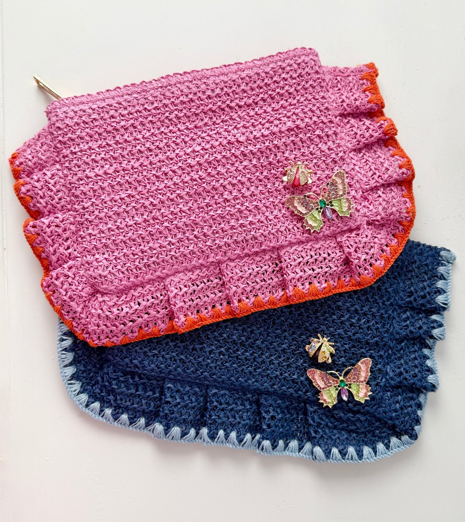 Woven straw clutch in Rosé pink and Capri blue with crystal butterfly and ladybug brooches pinned on front against a white background