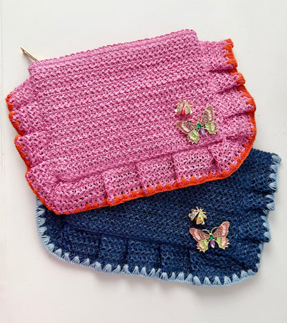 Woven straw clutch in Rosé pink and Capri blue with crystal butterfly and ladybug brooches pinned on front against a white background
