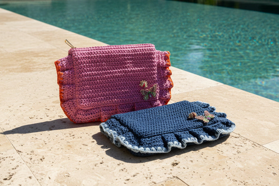 Two crocheted pouches, one pink and one blue, with butterfly embellishments next to a pool and outdoor setting.